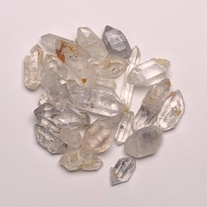 Könnte beinhalten: Eine Ansammlung klarer Quarz-Kristallspitzen, die in Größe und Form variieren, wobei einige interne Einschlüsse und subtile Farbvariationen aufweisen. Die Kristalle sind in einem losen Haufen angeordnet und zeigen ihre natürliche, rohe Schönheit.