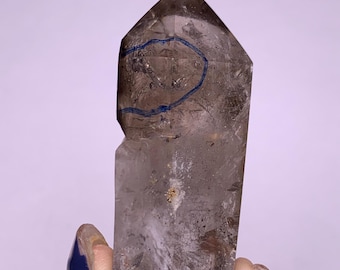 May include: A clear, smoky quartz crystal point with a faceted top. The crystal has a natural, rough texture and a blue line drawn on the surface. The crystal is held in a hand against a soft purple background.