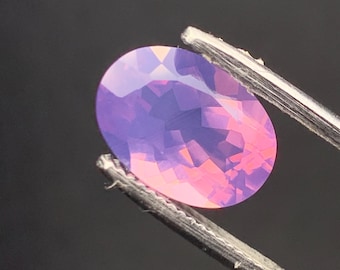 May include: An oval-shaped gemstone with a faceted surface, held by metal tweezers. The gem displays a gradient of colors, transitioning from pink to purple. The background is a solid black.