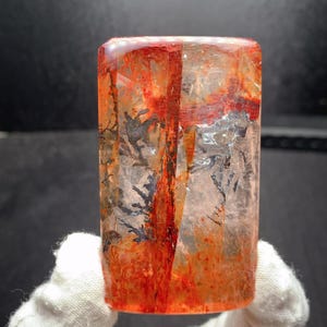 May include: A rectangular, translucent crystal with vibrant red and orange internal patterns. Dark, tree-like inclusions are visible within the crystal. The crystal is held by a gloved hand.