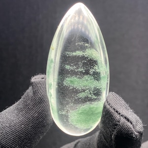 May include: A teardrop-shaped clear quartz gemstone featuring internal green mineral inclusions. The stone has a smooth, polished surface with a pointed top and a rounded base, showcasing natural green moss-like patterns suspended within the transparent crystal structure.