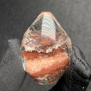 May include: A teardrop-shaped polished gemstone featuring a clear, transparent quartz exterior with prominent internal inclusions of deep red and earthy brown mineral deposits layered throughout the centre of the stone's structure.