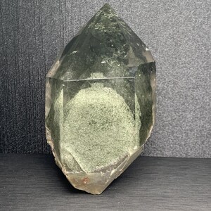 May include: A clear, pointed quartz crystal featuring a prominent internal phantom inclusion of dusty green mineral deposits. The crystal has a hexagonal structure with smooth, translucent faces and a textured, opaque green core visible inside.