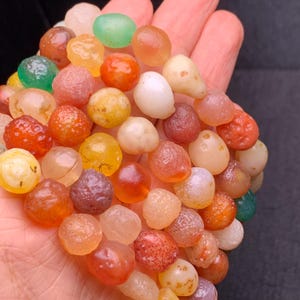 Puede incluir: Una hebra de cuentas de ágata de colores, que presenta una variedad de tonos de rojo, naranja, amarillo, verde y blanco. Las cuentas son lisas y pulidas, con un aspecto natural y orgánico.