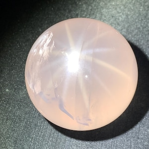 Puede incluir: Una piedra preciosa de cuarzo rosa esférica y lisa con una superficie pulida y translúcida. La piedra muestra un tono rosa pálido suave con inclusiones nubladas internas y un efecto de estrella de seis puntas visible.