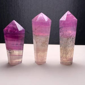 Puede incluir: Tres torres de cristal de fluorita pulidas. Los cristales presentan un degradado de colores, incluyendo morado, rosa y blanco. Los cristales tienen una parte superior facetada y una base rectangular. Los cristales se muestran sobre una superficie blanca.