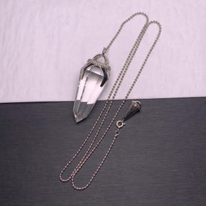 May include: A clear, faceted, pointed crystal pendant suspended from a silver-toned ball chain. The crystal is secured by a metallic cap featuring a textured, studded surface. A small, conical silver-toned metal weight is attached to the chain.