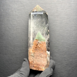 Pode incluir: Uma torre de cristal de quartzo transparente polido com uma inclusão interna proeminente, apresentando uma base texturizada castanho-avermelhada coberta por uma camada verde vibrante. O cristal possui uma ponta hexagonal e lados.