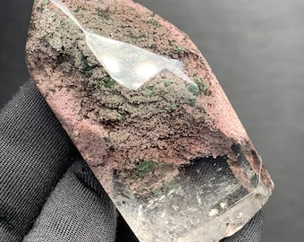 May include: A polished crystal point featuring a textured, dusty pink and brown upper section with embedded dark green mineral inclusions, transitioning into a clear, transparent base with visible internal fractures and light reflections.