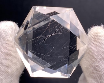 May include: A clear, faceted crystal with a geometric shape, held by a gloved hand. The crystal has internal inclusions and a polished surface, reflecting light. The crystal's facets create a multi-sided form, showcasing its clarity and intricate details.