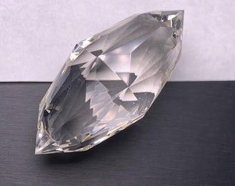 May include: A clear, faceted crystal gemstone with a pointed shape. The crystal has multiple geometric facets that reflect light, creating a sparkling effect. The gemstone is set against a black and white background.