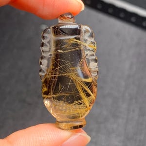 Puede incluir: Un colgante de cuarzo transparente tallado en forma de jarrón con inclusiones de rutilo dorado. El colgante tiene un diseño detallado con tallas intrincadas.