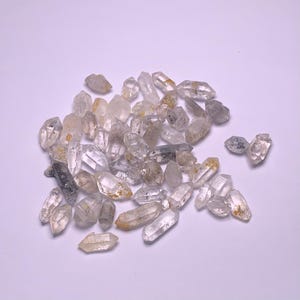 Pode incluir: Uma coleção de cristais de quartzo transparentes com uma aparência ligeiramente nublada. Os cristais são pontiagudos e têm um aspecto natural e bruto.