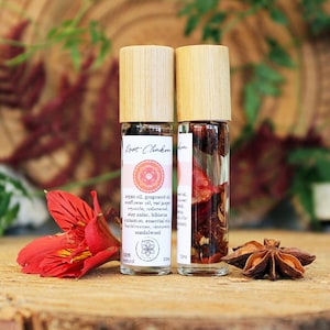 May include: Two glass roller bottles with bamboo lids filled with red and brown ingredients. The bottles are labeled "Root Chakra" and have a list of ingredients including argan oil, grapefruit oil, sunflower oil, red pepper crystals, sandalwood, and more. The bottles are sitting on a wooden surface with a red flower and a star anise.