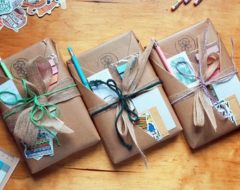 Può includere: Tre pacchi di carta marrone legati con spago e decorati con adesivi. Ogni pacco contiene un quaderno, una matita e altri articoli di cancelleria.