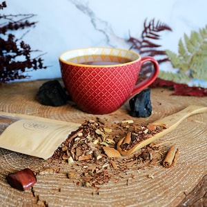 Op de afbeelding: Een rode theekop gevuld met donker amberkleurige thee staat op een houten blok met een bruine papieren zak en een houten lepel gevuld met losse thee. De theebladeren zijn een mix van bruin, rood en beige.