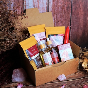 Pode incluir: Uma caixa de cartão castanha cheia de produtos de autocuidado. A caixa contém pequenos frascos de óleo, saquetas, uma vela vermelha e um baralho de cartas com a etiqueta "Sexual Magic Tarot". Cristais de quartzo rosa e pétalas de rosa secas estão espalhadas.
