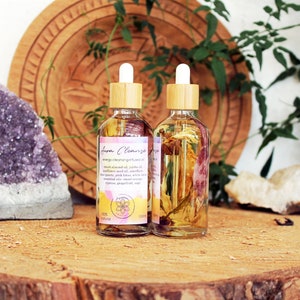 Peut inclure: Deux flacons en verre avec des compte-gouttes en bambou remplis d'un liquide clair et de fleurs séchées. Les flacons portent des étiquettes indiquant "Aura Cleanse Energy Cleansing Oil" avec une liste d'ingrédients. Les flacons sont posés sur une surface en bois avec un cristal d'améthyste violet et une assiette en bois en arrière-plan.