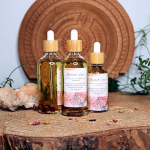 以下が含まれることがあります： 木製の表面に、「Breast Love」と書かれたラベルが付いた、竹製のスポイトと琥珀色のガラス瓶が3本あります。瓶には透明な液体が入っており、乾燥したバラの花びらとピンク色のクリスタルで囲まれています。