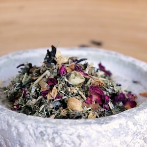 Könnte beinhalten: Eine Nahaufnahme einer weißen Keramikschale, gefüllt mit einer Mischung aus getrockneten Kräutern, Blüten und Gewürzen. Die Mischung enthält grüne Kräuter, getrocknete Rosenblätter und kleine braune Stücke.