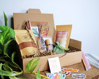 May include: A cardboard box filled with a variety of items, including a journal, a pencil, a small bag of brown powder, a bottle of essential oil, a small green bag, a bag of bath salts, and a set of stickers. The box is sitting on a wooden surface.