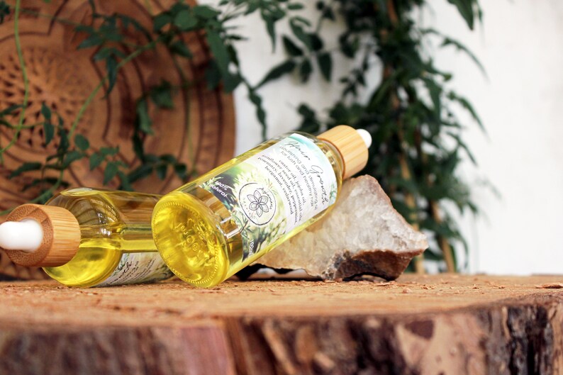 Puede incluir: Dos botellas de vidrio &aacute;mbar con goteros y tapas de madera, llenas de un l&iacute;quido amarillo. Las botellas est&aacute;n etiquetadas con "Garden Goodness" y tienen un dise&ntilde;o floral. Las botellas est&aacute;n sobre una superficie de madera con una roca y follaje verde en el fondo.