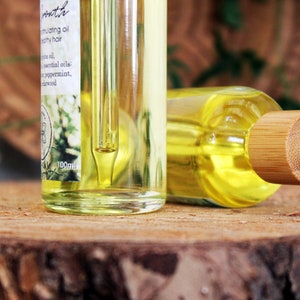 Puede incluir: Dos botellas de vidrio transparente con goteros, llenas de un l&iacute;quido amarillo. Las botellas est&aacute;n sobre una superficie de madera. La botella de la izquierda tiene una etiqueta que dice "Promote Nourishing oil for healthy hair Jojoba oil, essential oils: Tea tree, peppermint, sandalwood 100ml". La botella de la derecha tiene una tapa de bamb&uacute; con una punta de gotero blanca.