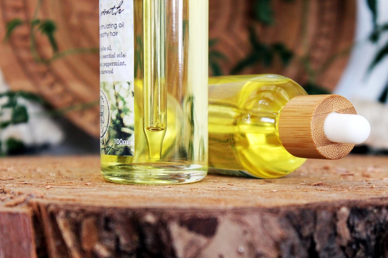 Puede incluir: Dos botellas de vidrio transparente con goteros, llenas de un l&iacute;quido amarillo. Las botellas est&aacute;n sobre una superficie de madera. La botella de la izquierda tiene una etiqueta que dice "Promote Nourishing oil for healthy hair Jojoba oil, essential oils: Tea tree, peppermint, sandalwood 100ml". La botella de la derecha tiene una tapa de bamb&uacute; con una punta de gotero blanca.
