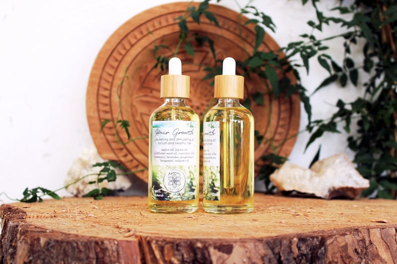 Puede incluir: Dos botellas de vidrio &aacute;mbar con goteros de bamb&uacute; llenas de un l&iacute;quido transparente. Las botellas est&aacute;n etiquetadas con "Hair Growth" y contienen una mezcla de aceites para nutrir y estimular el crecimiento del cabello. Las botellas est&aacute;n sobre una superficie de madera con follaje verde en el fondo.