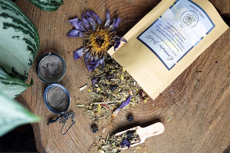 May include: A brown paper bag with the text "Dream Well" and "100% Natural" printed on it. The bag contains a loose blend of dried herbs and flowers, including blue and purple petals, green leaves, and small black berries. There are two metal tea infusers next to the bag, one open and one closed.