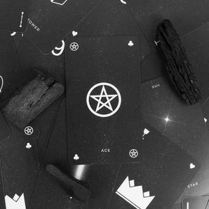May include: Black and white tarot cards with various symbols and text, including "Ace", "Tower", "Temperance", "King", and "Star". The Ace card features a pentagram. Two pieces of dark wood are placed on the cards.