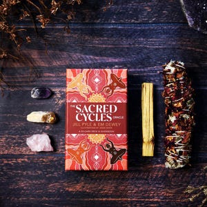 May include: A red and orange oracle card deck titled "The Sacred Cycles" with a 50-card deck and guidebook. The image also includes crystals, a palo santo stick, and a bundle of dried herbs on a dark wooden surface.