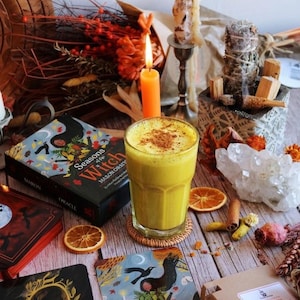 Puede incluir: Un vaso de latte de cúrcuma dorado espolvoreado con canela, rodeado por un mazo de oráculo Seasons of the Witch, una vela naranja encendida, un grupo de cristales, rodajas de naranja seca, un atado de salvia y cartas tituladas Darkness Falls y Falling Leaves.