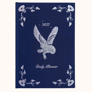 Könnte beinhalten: Ein dunkelblauer Tagesplaner mit einer silbernen Folienillustration einer fliegenden Eule in der Mitte. Der Text 2027 und Daily Planner sind in Silber gedruckt. Die Ecken zeigen silberne florale Rankenmotive mit kleinen Sternen.