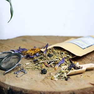 May include: A brown paper bag filled with loose leaf herbal tea blend. The tea blend includes dried flowers, herbs, and berries. There are two metal tea infusers and a wooden scoop on a wooden surface.