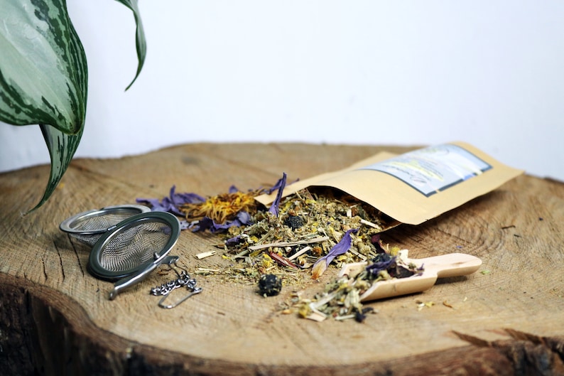 May include: A brown paper bag filled with loose leaf herbal tea blend. The tea blend includes dried flowers, herbs, and berries. There are two metal tea infusers and a wooden scoop on a wooden surface.
