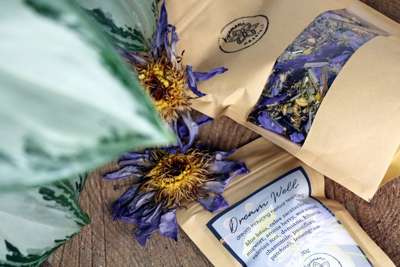 May include: A brown paper bag with a clear window showing a blend of dried herbs and flowers. The bag is labeled "Dream Well" and lists the ingredients: blue lotus, calea zacatechichi, mugwort, aronia berry, valerian root, damiana, hibiscus, chamomile, passiflora, patchouli, and lemongrass. The bag is 30g.