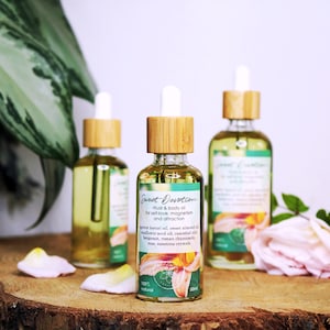 以下が含まれることがあります： 竹製のスポイトと「Sweet Devotion Ritual & Body Oil for self-love, magnetism and attraction」と書かれたラベルが付いた琥珀色のガラス瓶が3本。瓶は、ピンクのバラの花びらが置かれた木製の表面の上に置かれています。