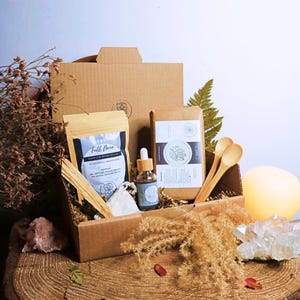 Puede incluir: Una caja de cartón marrón llena de una variedad de artículos, que incluyen una bolsa de polvo para baño y cuerpo "Full Moon", una pequeña botella de aceite, dos cucharas de madera y un trozo de tela blanca. La caja está sentada sobre una superficie de madera con flores secas y un cristal.
