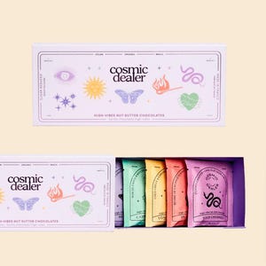 Op de afbeelding: Een witte doos met "Cosmic Dealer" high-vibes notenboter chocolade. De doos is versierd met kleurrijke illustraties en tekst. Binnenin zijn individuele chocoladerepen in verschillende kleuren zichtbaar.