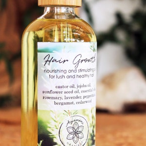 Puede incluir: Una botella de vidrio transparente con una tapa de madera, etiquetada como "Hair Growth" y que enumera ingredientes como aceite de ricino, aceite de jojoba, aceite de semilla de girasol, romero, lavanda, menta piperita, bergamota y madera de cedro.