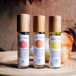 Może przedstawiać: Trzy małe szklane butelki z bambusowymi korkami i etykietami. Etykiety są białe z czerwonymi, pomarańczowymi i żółtymi kółkami i tekstem, który brzmi "Root Chakra", "Sacral Chakra" i "Solar Plexus". Butelki są wypełnione przezroczystym płynem i stoją na drewnianej powierzchni.