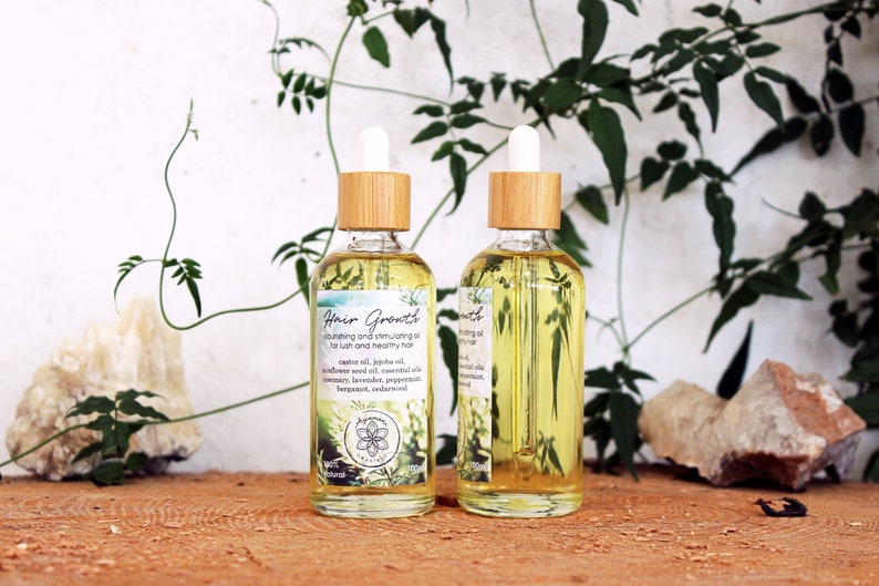Puede incluir: Dos botellas de vidrio &aacute;mbar con goteros de bamb&uacute; y etiquetas que dicen "Hair Growth" en escritura blanca. Las botellas est&aacute;n llenas de un l&iacute;quido transparente y amarillo y est&aacute;n sobre una superficie de madera. Las botellas est&aacute;n rodeadas de follaje verde y rocas.