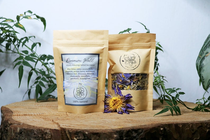May include: Two brown paper bags with a clear window, containing dried herbs and flowers. The bags are labeled "Dream Well" and list the ingredients, including blue lotus, valerian root, mugwort, and chamomile. The text "100% natural" is printed on the bag.