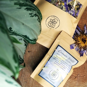 May include: A brown paper bag with a label that reads "Dream Woll dream inducing herbal tea" and lists ingredients including blue lotus, calea zacatechichi, mugwort, aronia berry, valerian root, damiana, chamomile, patchouli, and lemon balm. The bag is sitting on a wooden surface with a dried flower.