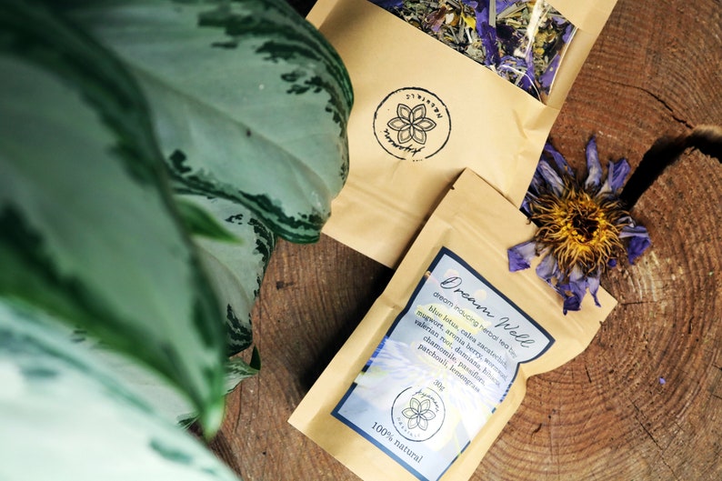 May include: A brown paper bag with a label that reads "Dream Woll dream inducing herbal tea" and lists ingredients including blue lotus, calea zacatechichi, mugwort, aronia berry, valerian root, damiana, chamomile, patchouli, and lemon balm. The bag is sitting on a wooden surface with a dried flower.