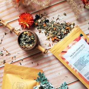 May include: Overhead view of herbal tea and packaging. A wooden scoop holds loose tea, with more scattered on a wooden surface. Two tea packages are visible, one labelled "Sacral Chakra Tea" and the other with a floral logo. Dried flowers and foliage are arranged around the tea.