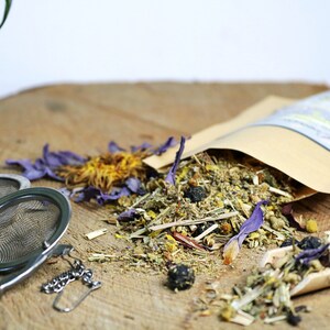 May include: A brown paper bag filled with loose leaf herbal tea blend. The tea includes dried flowers, herbs, and berries. The bag is open and the tea is spilling out onto a wooden surface. There are two tea infusers next to the bag.