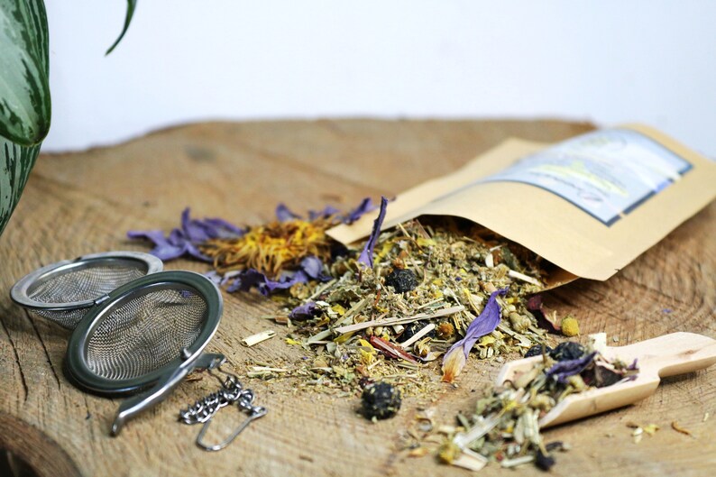 May include: A brown paper bag filled with loose leaf herbal tea blend. The tea includes dried flowers, herbs, and berries. The bag is open and the tea is spilling out onto a wooden surface. There are two tea infusers next to the bag.