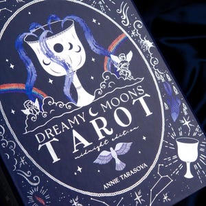 May include: A dark blue rectangular tarot deck box titled Dreamy Moons Tarot Midnight Edition by Annie Tarasova. The cover features white and blue illustrations of a chalice, cherubs, rainbows, stars, constellations, and a bird in flight.